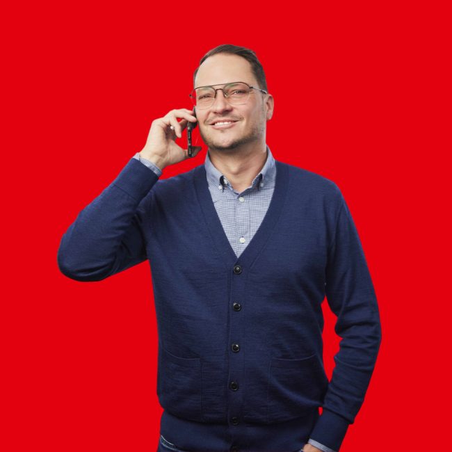 Franklyn-Wolf Mann in dunkelblauer Weste und hellblauer Bluse mit Telefon in der Hand